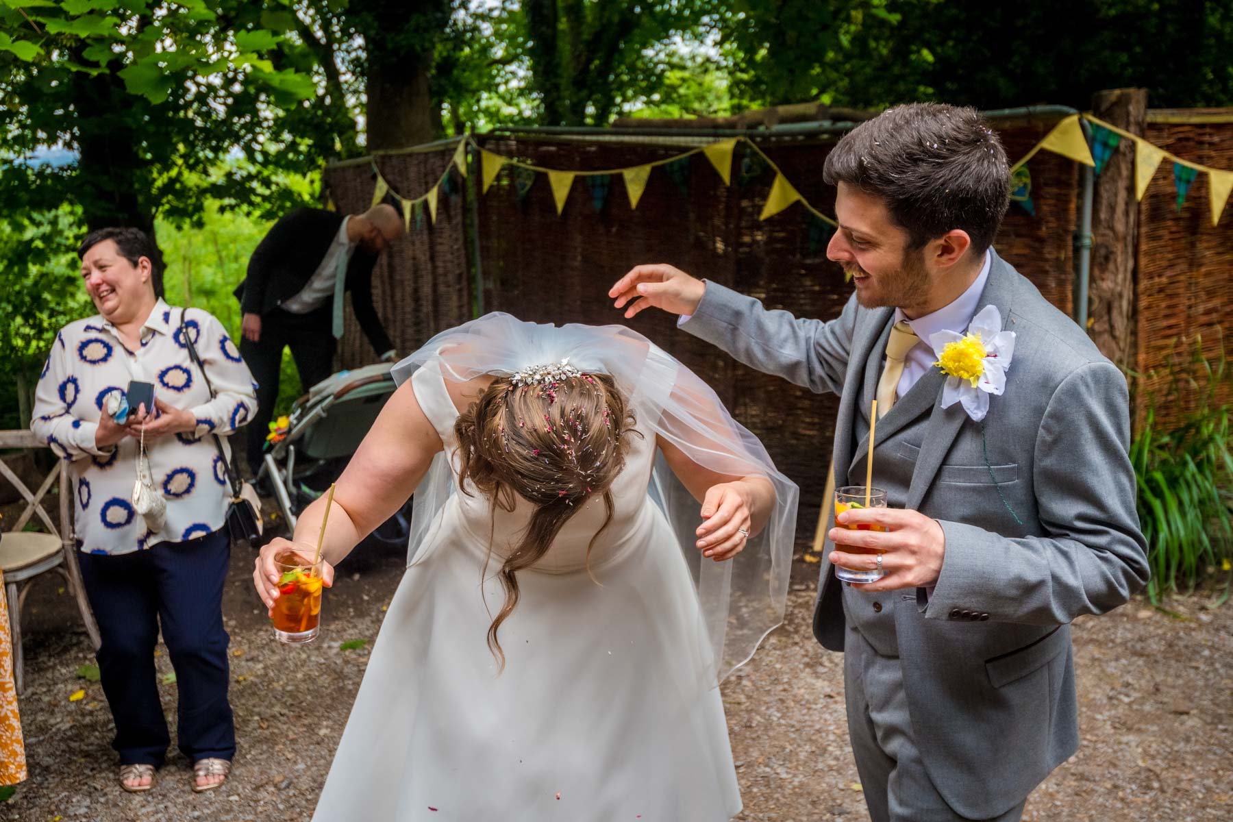 Bride bending over as laughing groom picks confetti out of her hair.