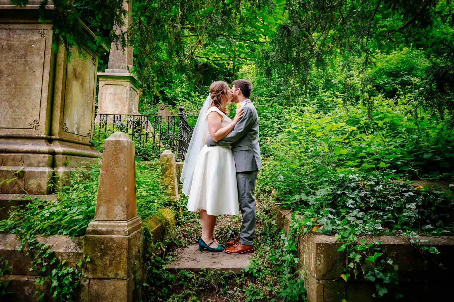 Bride kissing groom on cheek next to gravestones in Arnos Vale Cemetery.