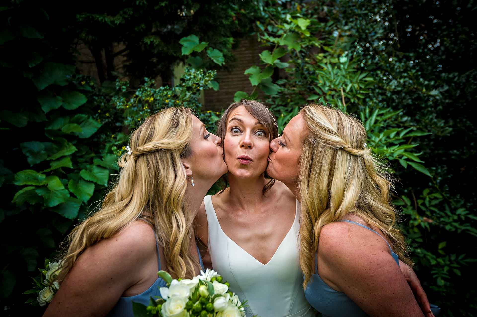 Fun Portrait of Bride Being Kissed on Cheek by Friends at Wedding